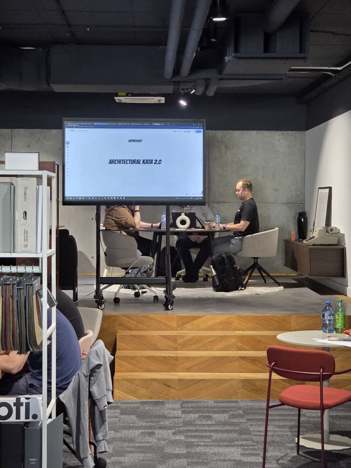 Zespół pracuje przy stole, na ekranie plansza Miro z napisem Architectural KATA 2.0